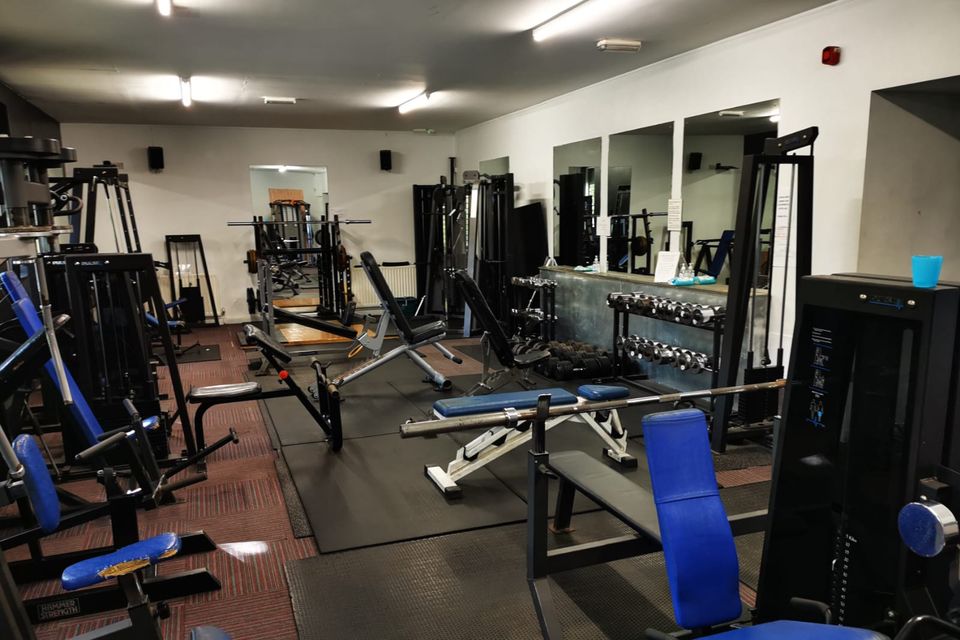 The main gym floor, with resistance machines down one side, benches on rubber matting, racks of dumbbells and mirrors along the wall.
